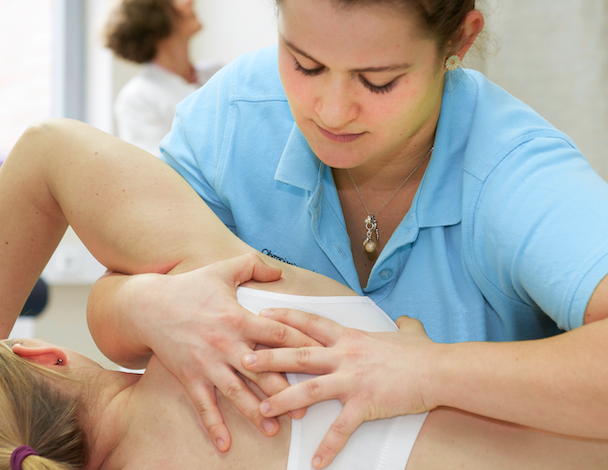 Therapeutin führt eine manuelle Therapie an der Schulter einer liegenden Patientin durch.